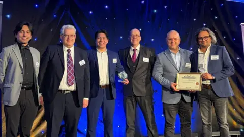 Университет Эссекса. Группа мужчин в строгих рубашках и блейзерах улыбается, держа в руках сертификат в рамке