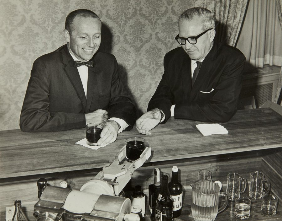 Black and white photo of two men at a bar being served drinks by a robotic arm