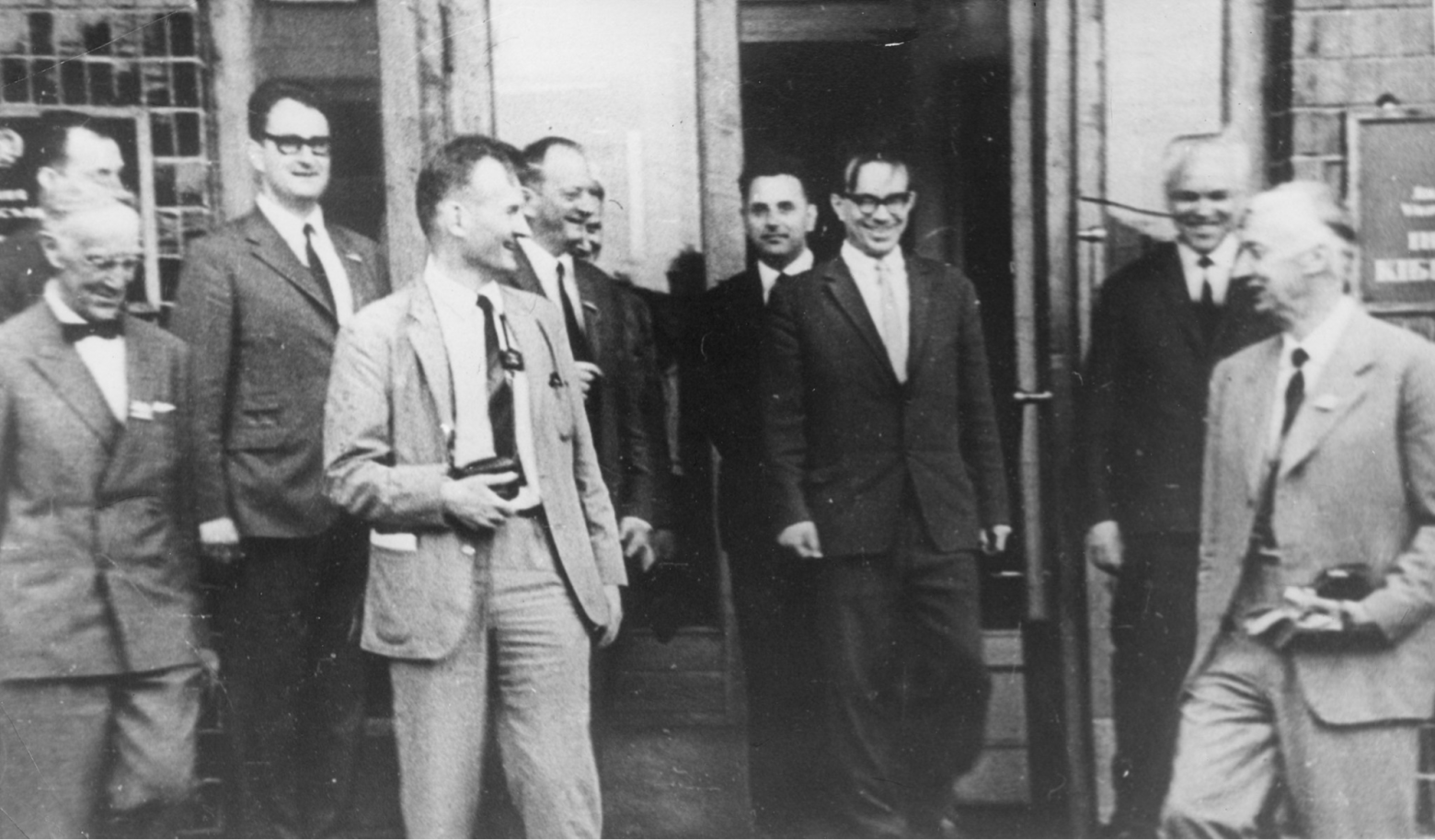 Виктор Глушков — в темном костюме в дверном проеме, в очках.
